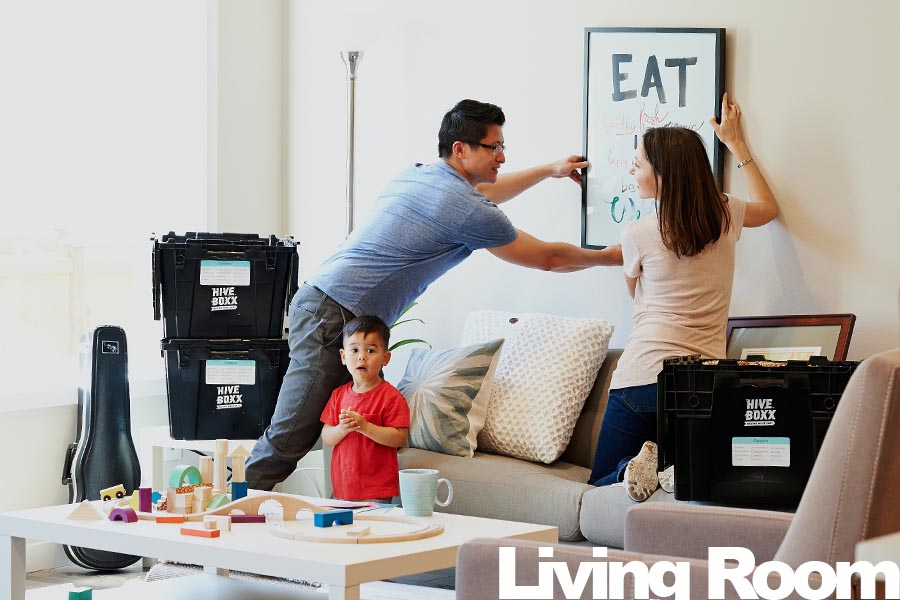 A family of three hanging a framed artwork on a wall in a living room with moving boxes nearby.