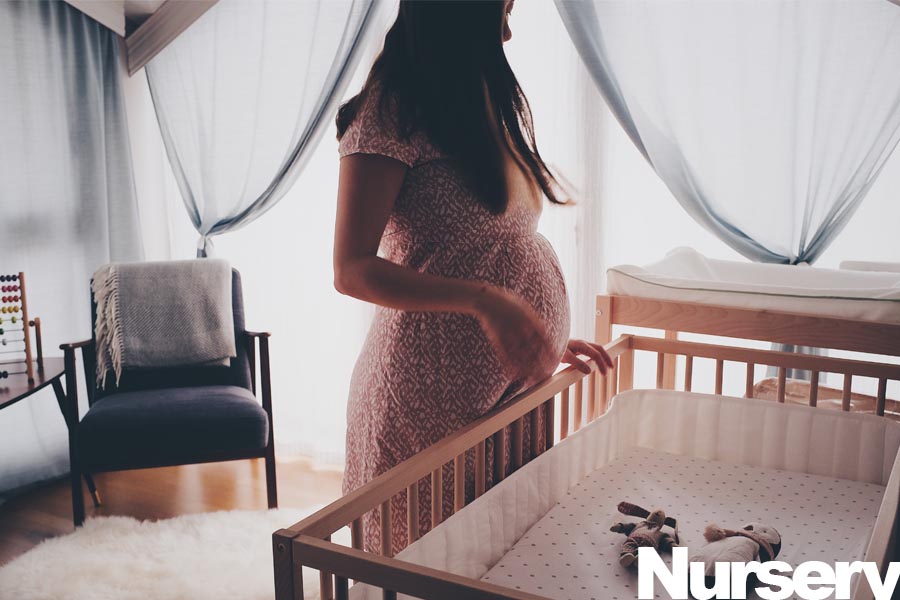 Pregnant woman in a patterned dress standing beside a wooden crib in a nursery.