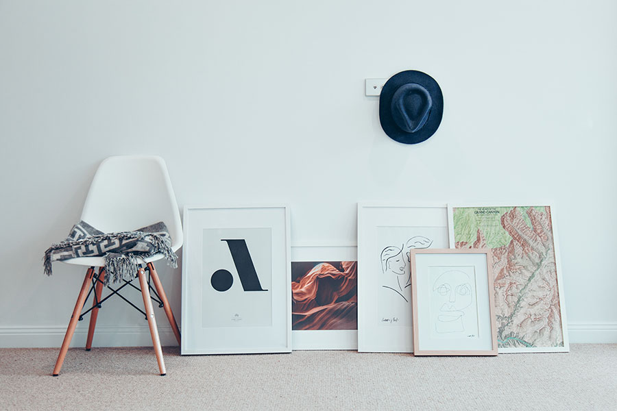 A collection of framed art prints leaning against a wall next to a chair and a hat.