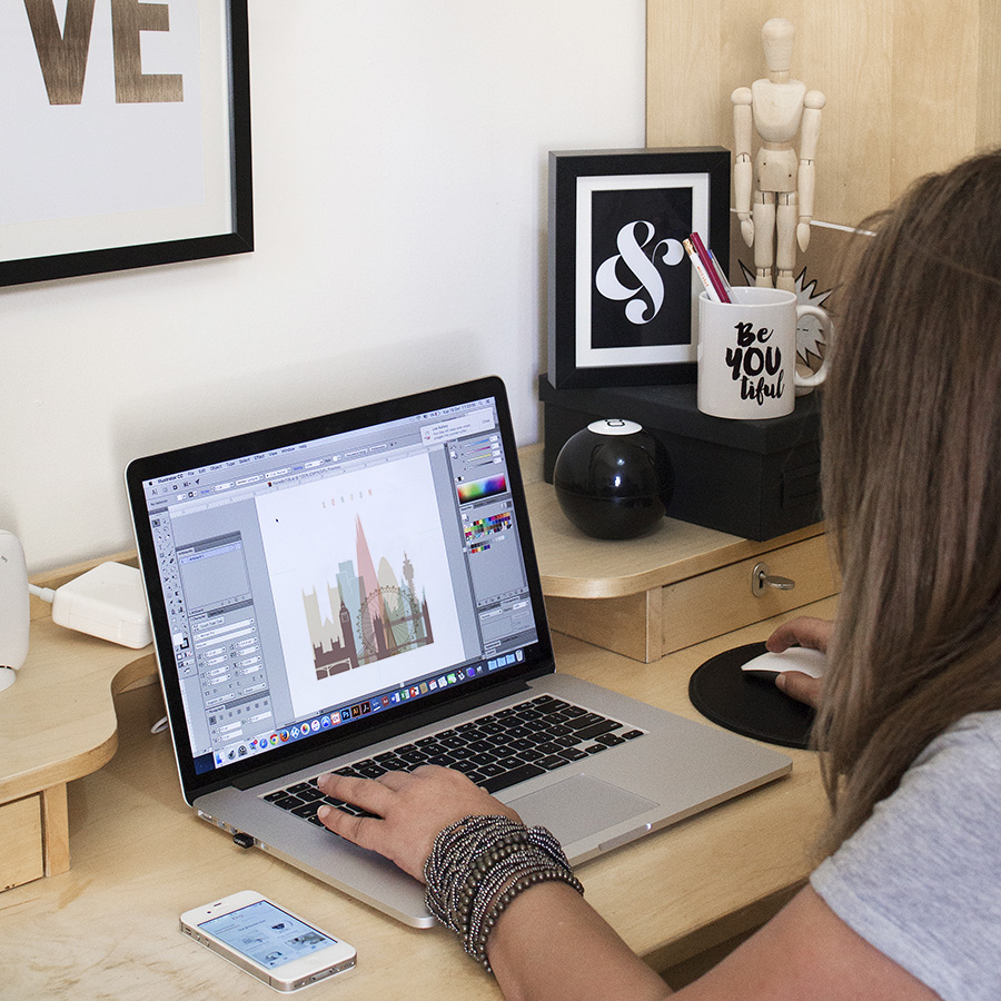 Vicky on "action" using a laptop while designing graphics at a wooden desk with various items.